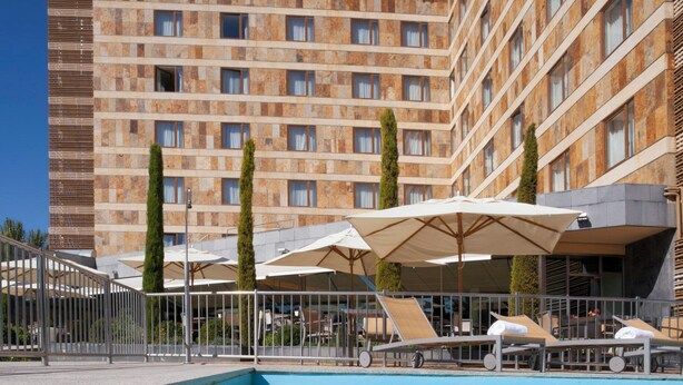 Une piscine entourée de chaises longues et de parasols, elle-même délimitée par une clôture, avec un immeuble à plusieurs étages en arrière-plan.
