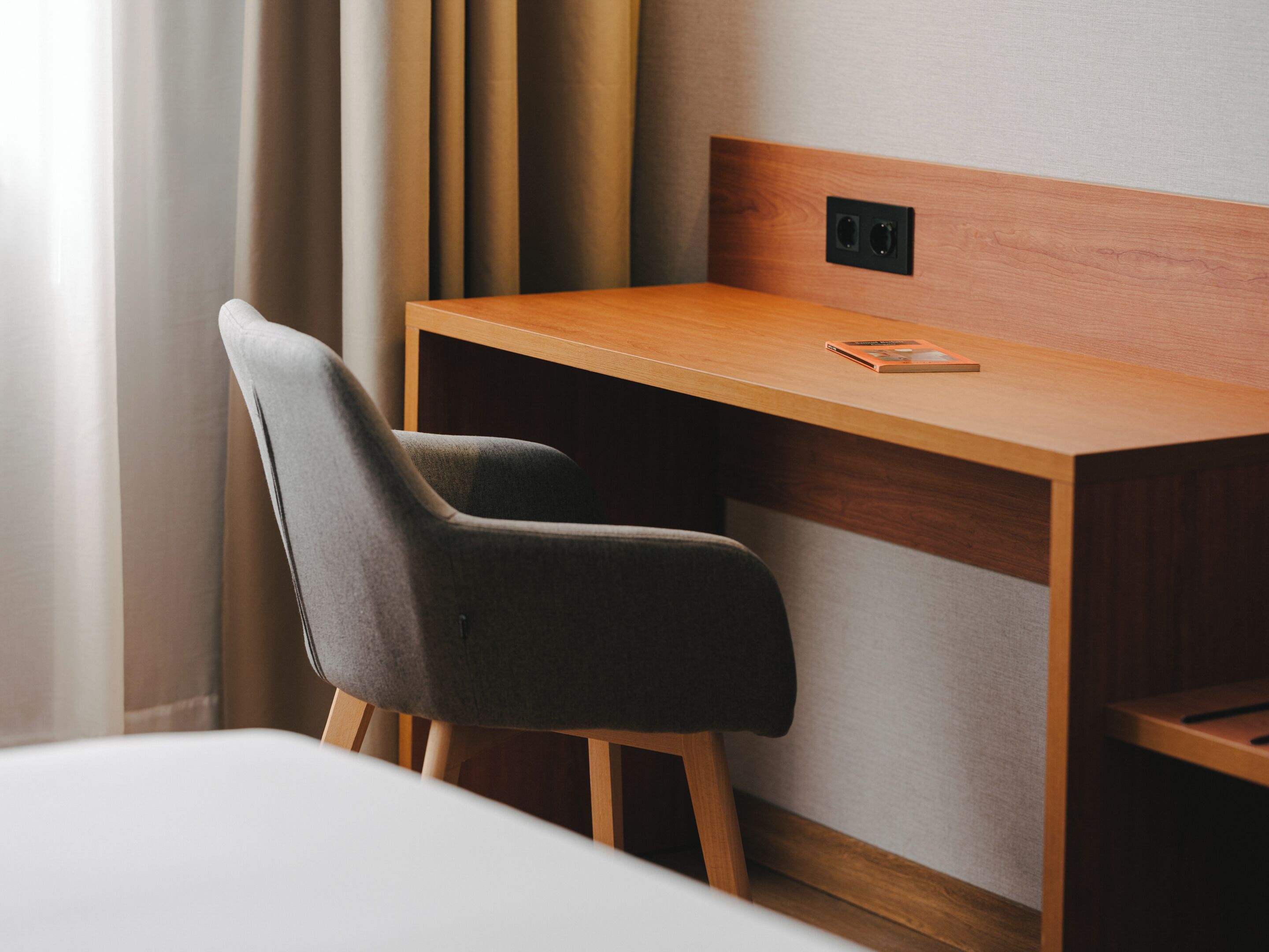 A modern hotel room with a wooden desk, a grey upholstered chair, and a white bed.