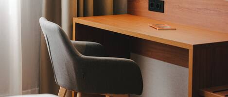 A modern hotel room with a wooden desk, a grey upholstered chair, and a white bed.