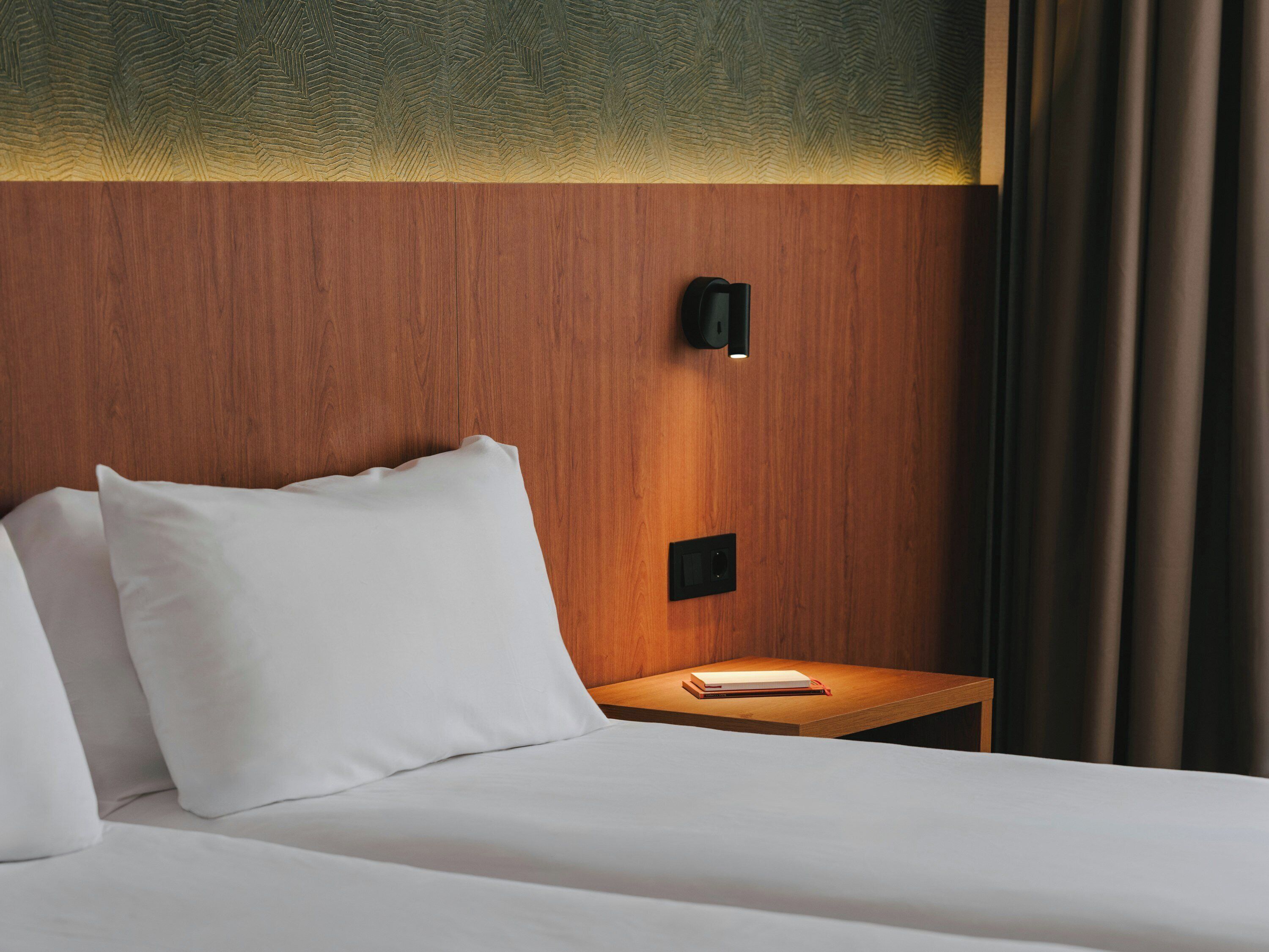 Ein Hotelzimmer mit einem Holzkopfteil, einem Nachttisch mit einem Buch und einer Wandleuchte.