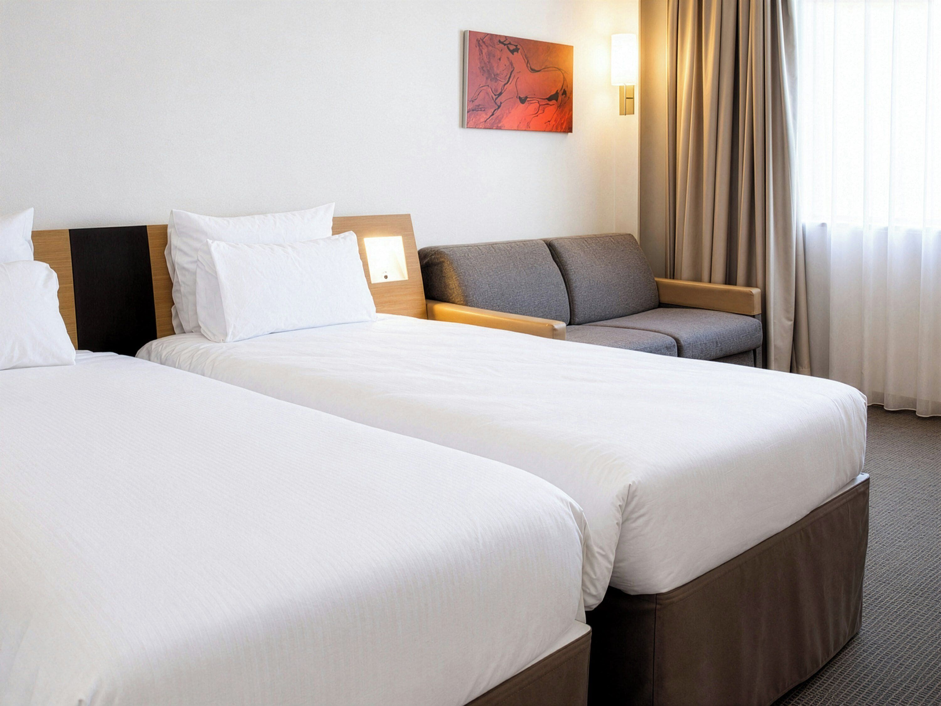 Habitación de hotel con dos camas, un sofá, un cuadro en la pared y una ventana con cortinas.