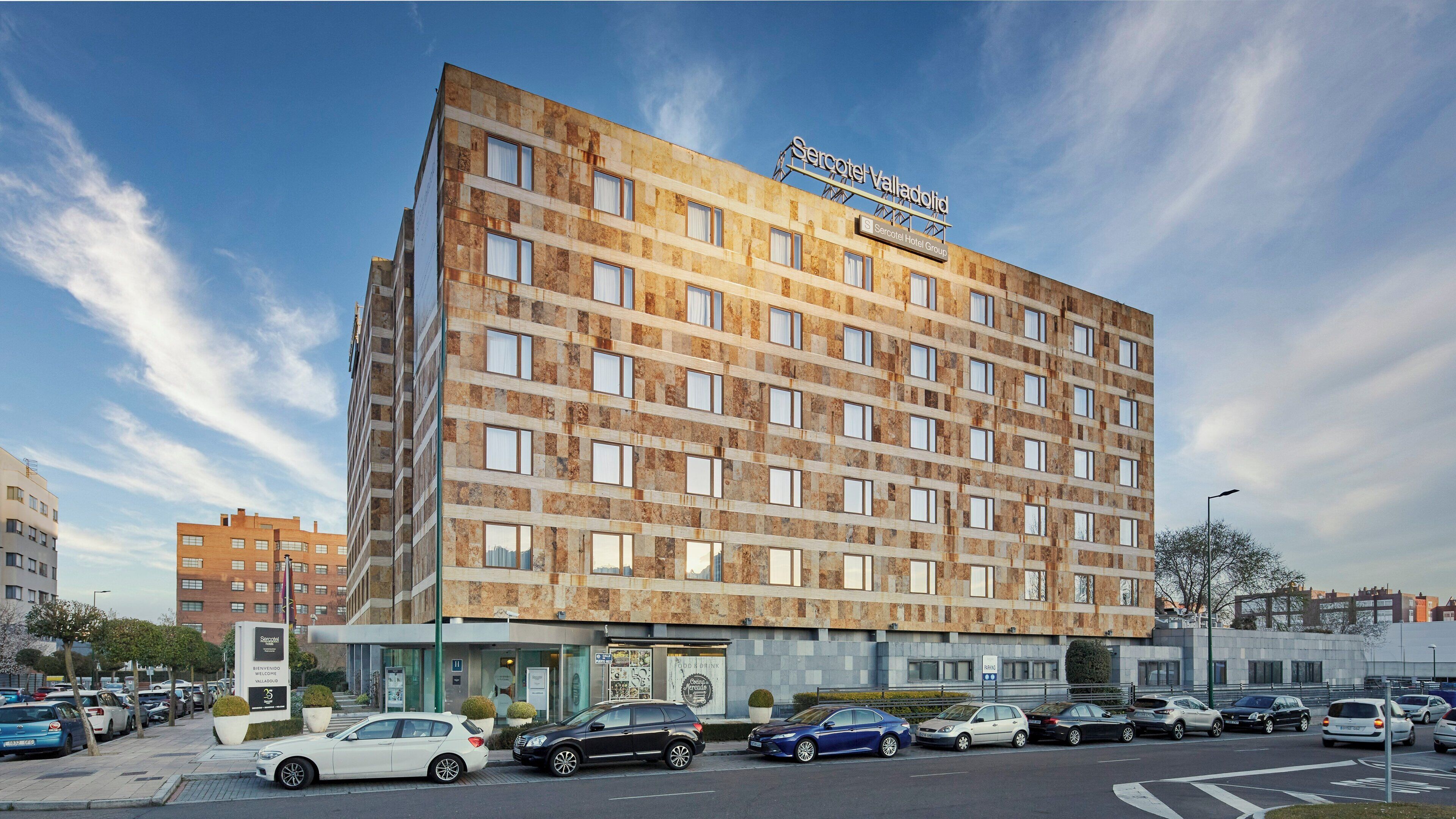 Um hotel de vários andares com fachada de pedra, placa visível do Serco Hotel Group e um estacionamento com vários carros.