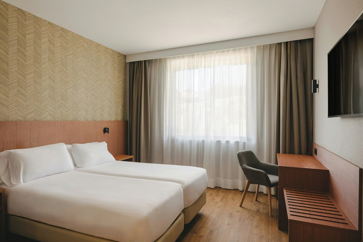 A hotel room with two beds, a desk, a chair, a TV, and a window with curtains.