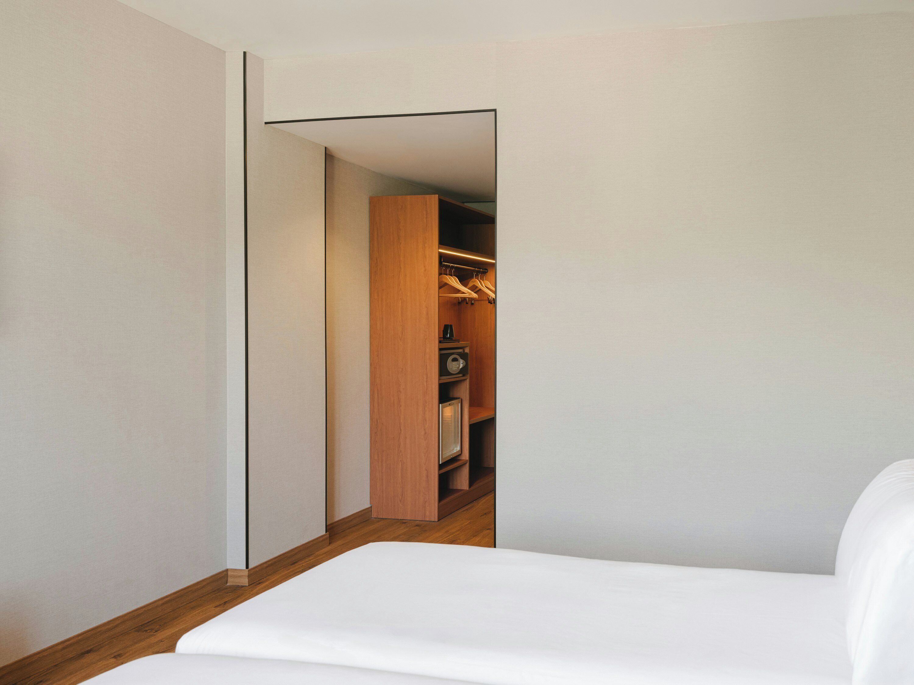 A bedroom with a wooden floor, a white bed, and a closet with hanging clothes.
