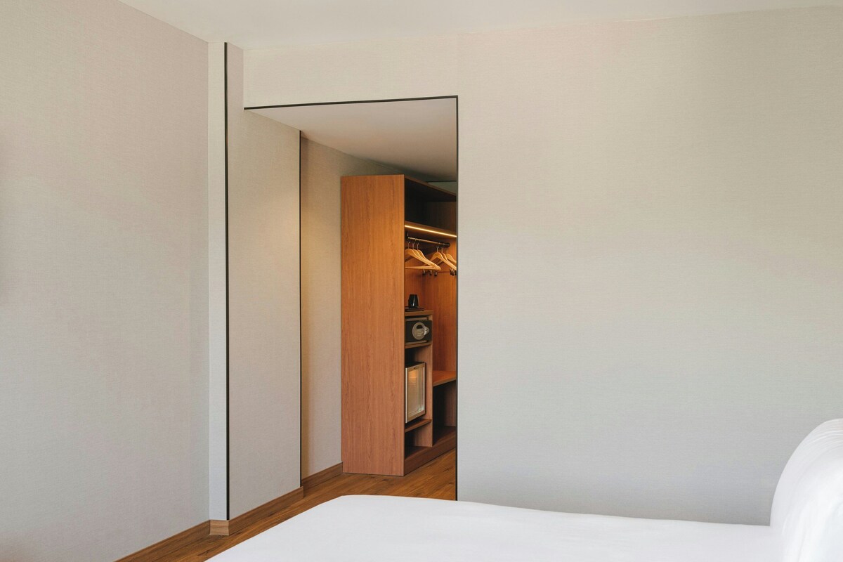A bedroom with a wooden floor, a white bed, and a closet with hanging clothes.