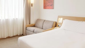 A hotel room with a bed, a sofa, a bedside table, a lamp, and a painting on the wall.