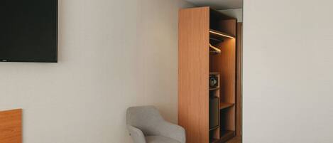 A modern hotel room with a bed, a grey armchair, a wooden wardrobe, and a flat-screen TV mounted on the wall.