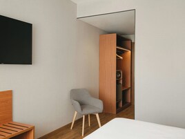 Una habitación de hotel moderna con una cama, un sillón gris, un armario de madera y un televisor de pantalla plana montado en la pared.