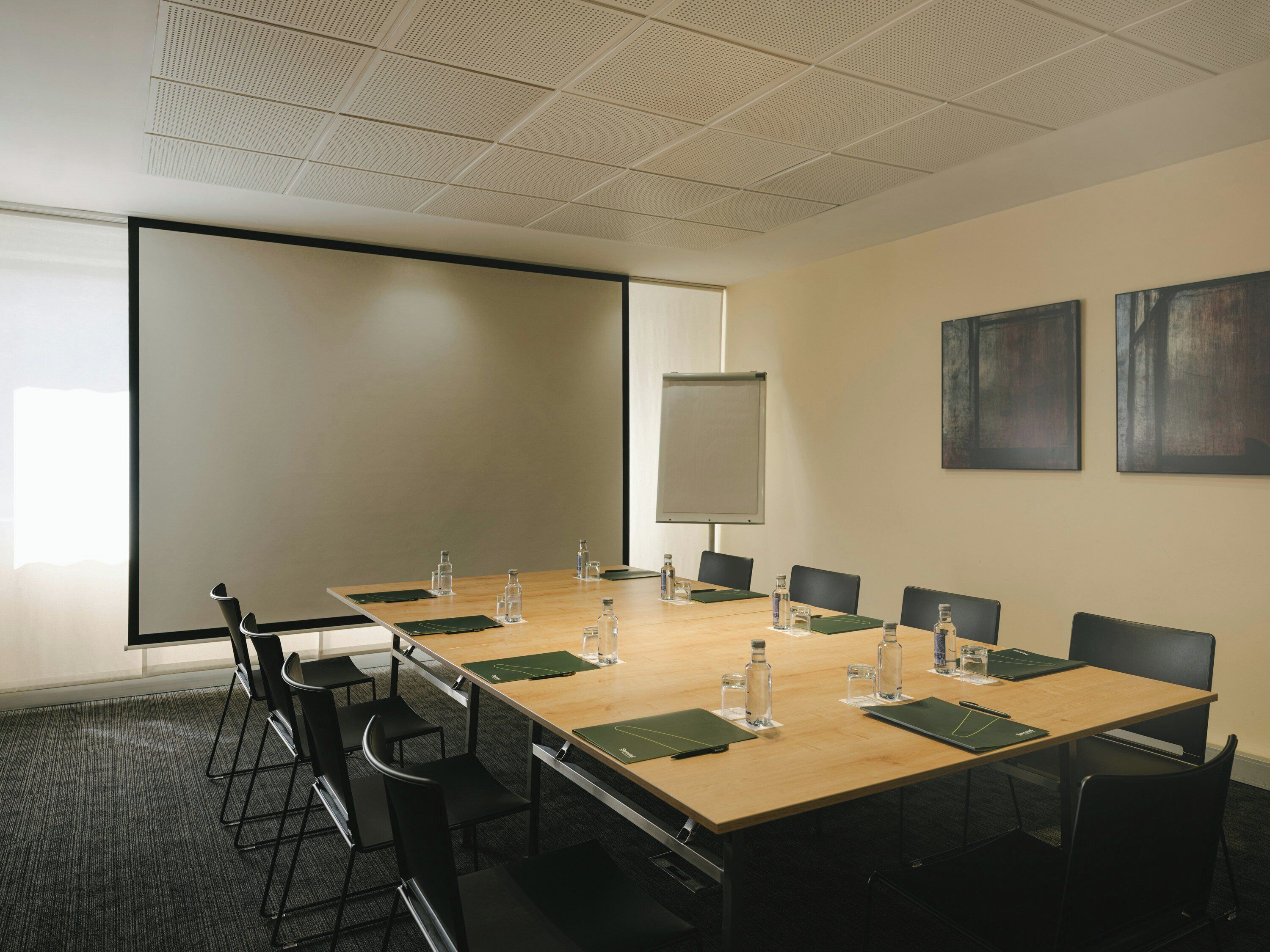 A conference room with a large table, chairs, a projector screen, and two abstract paintings on the wall.