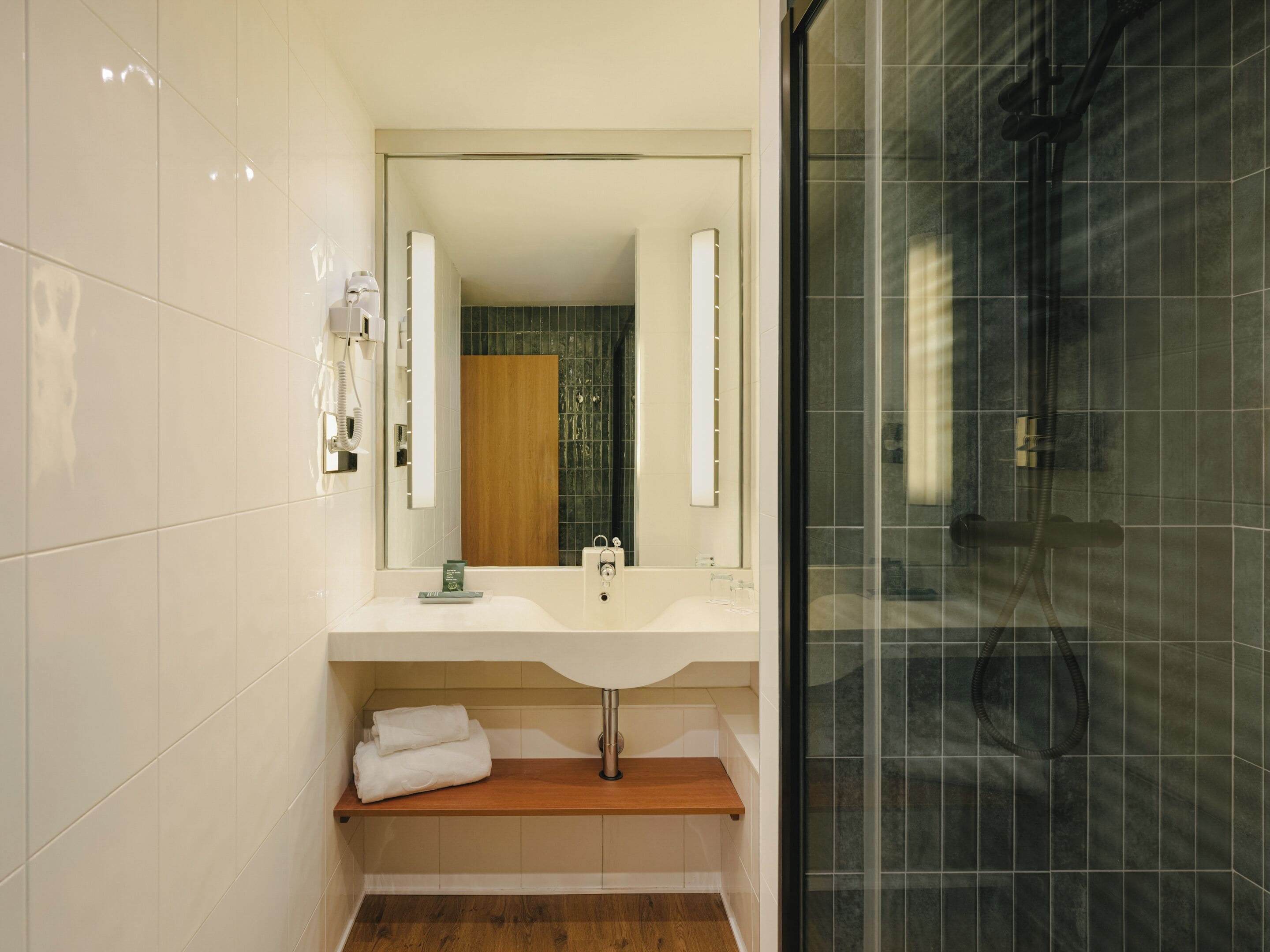 Un baño moderno con lavamanos blanco, repisa de madera, espejo grande y cabina de ducha con vidrio.