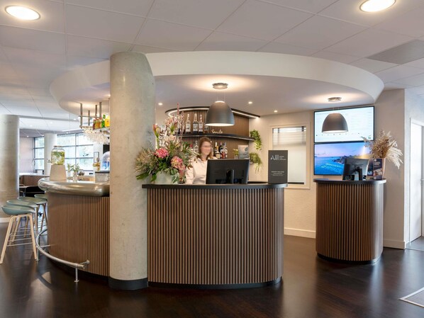 A reception area with a curved counter, a large pillar, and a monitor displaying a scenic view. - Novotel Suites Nancy Centre (Nancy)