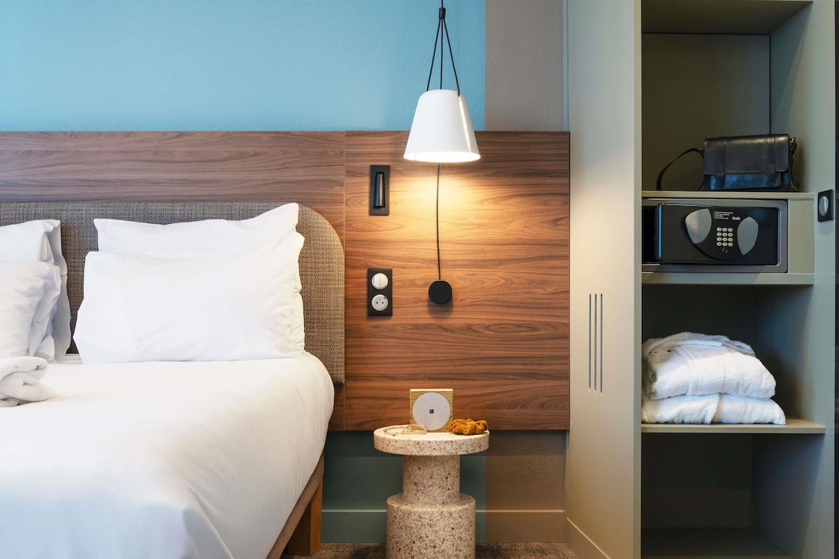 A hotel room with a bed, a nightstand with a lamp, a small table with slippers, and a wardrobe with towels.