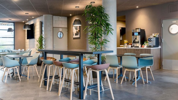 Un café moderno con una mesa central rodeada de sillas coloridas, una planta grande y una máquina de café.