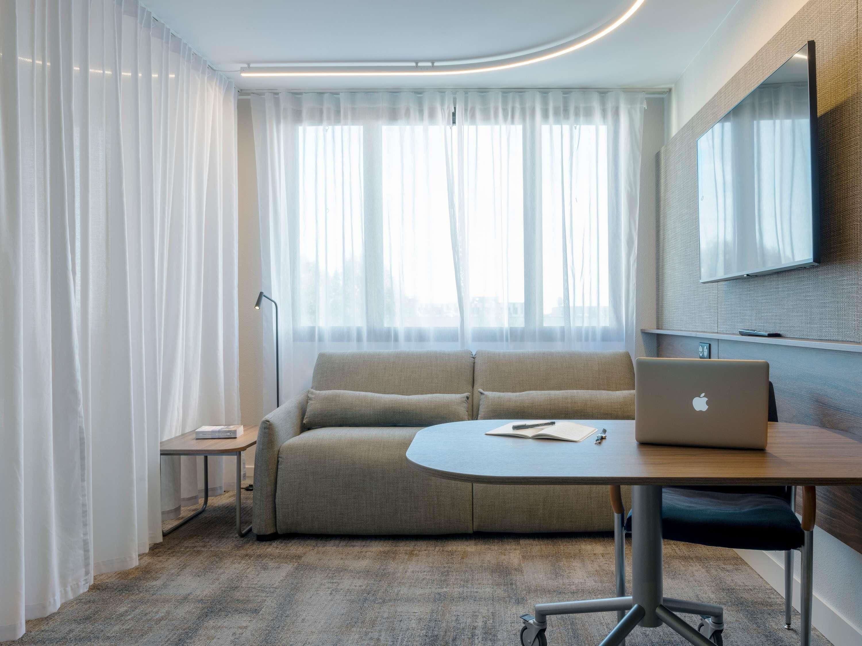 A modern hotel room with a sofa, a small table with an apple laptop, a floor lamp, and a TV mounted on the wall.