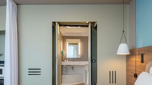Una habitación de hotel con cama, lámpara de noche, baño con espejo y piso de madera.