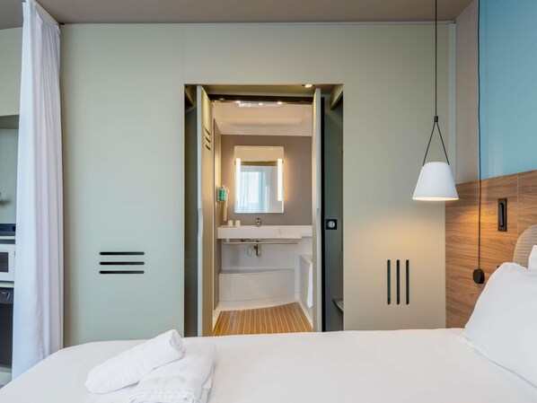 Una habitación de hotel con cama, lámpara de noche, baño con espejo y piso de madera.