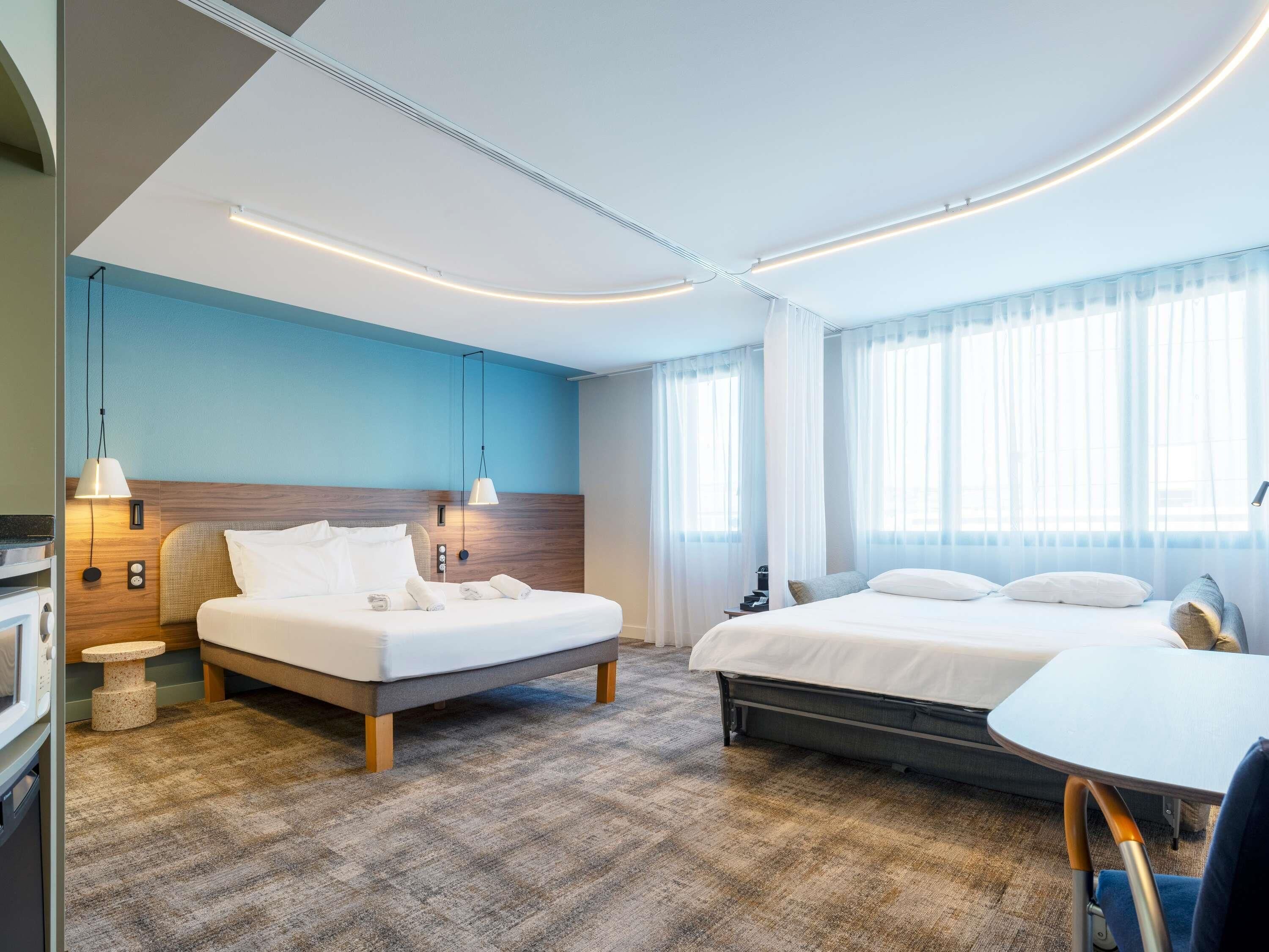 A modern hotel room with two beds, a small table, and a view of the city.