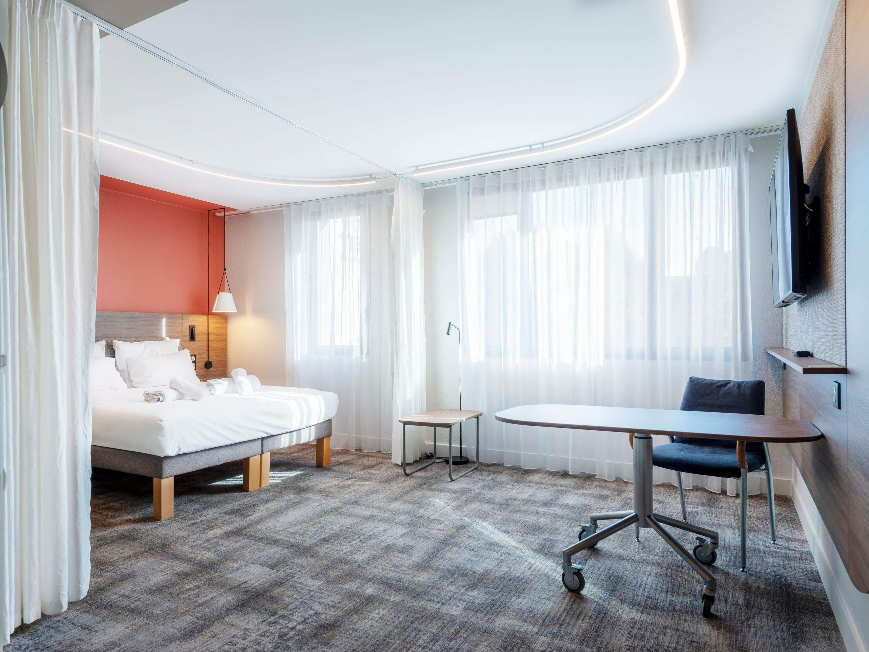Una habitación de hotel con cama, escritorio, silla, televisión y ventana con cortinas.