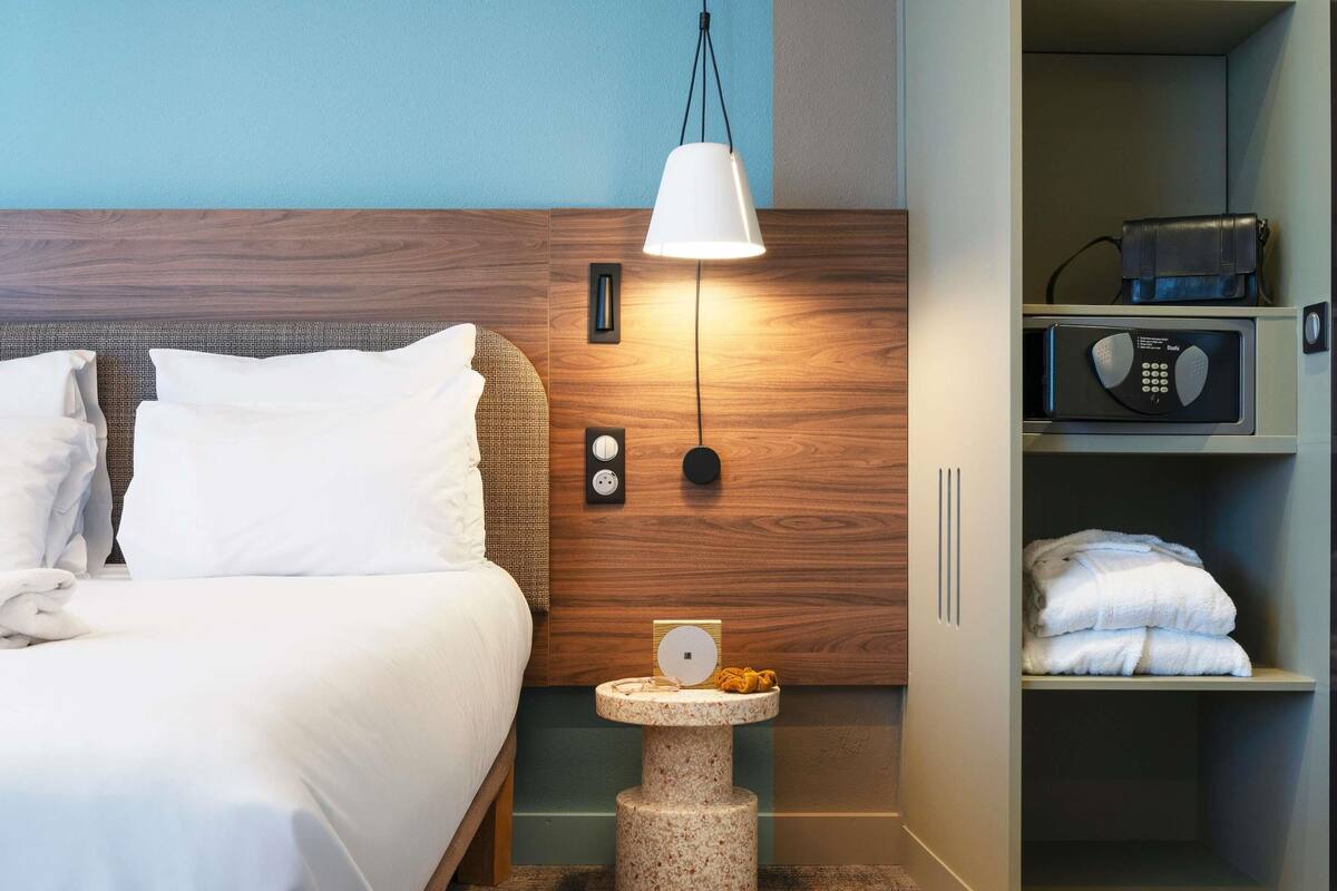Una habitación de hotel con una cama, una mesita de noche con lámpara, una pequeña mesa con zapatillas y un armario con toallas.