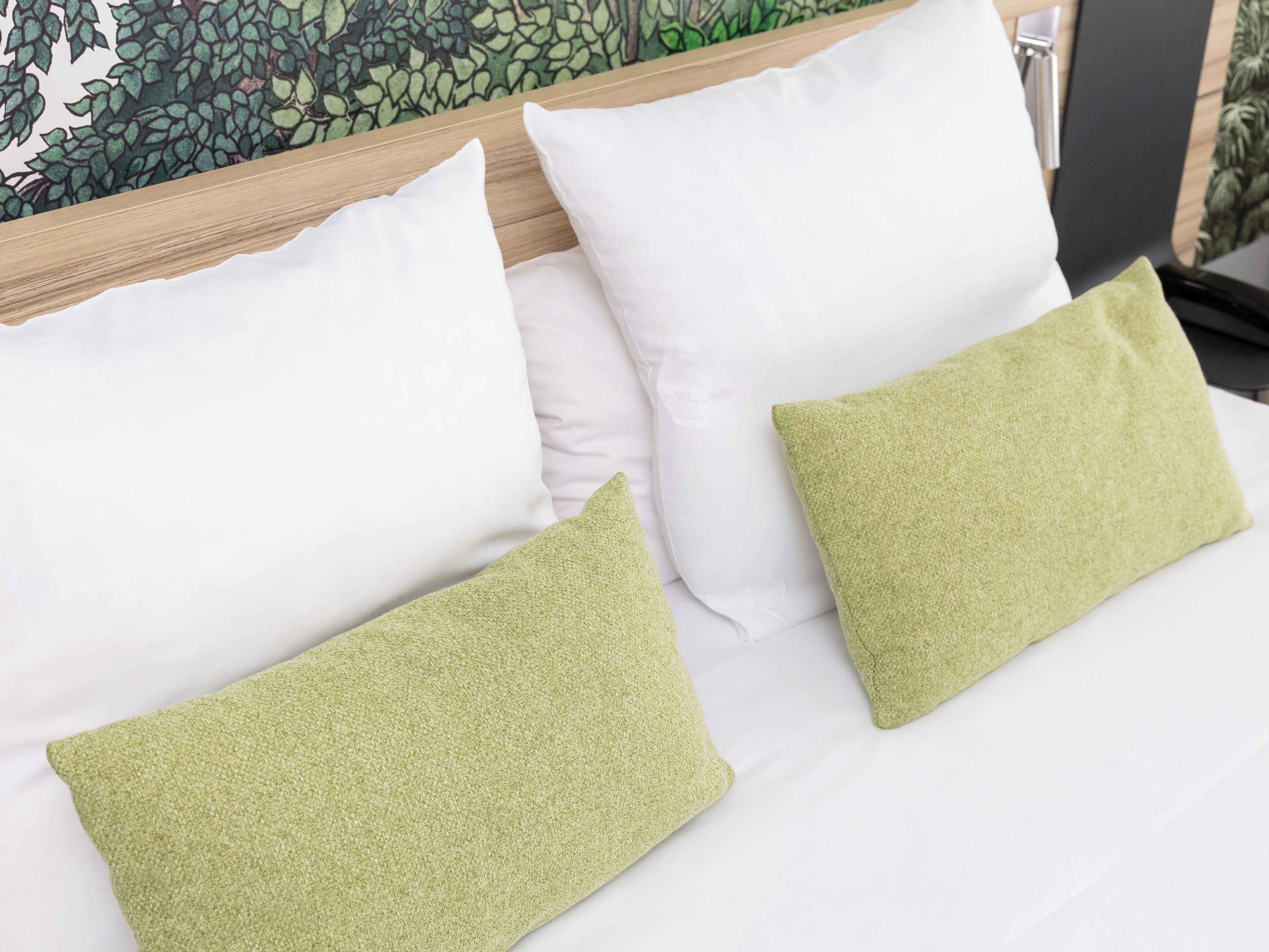 A bed with white and green pillows, a wooden headboard, and a green leafy wall mural.
