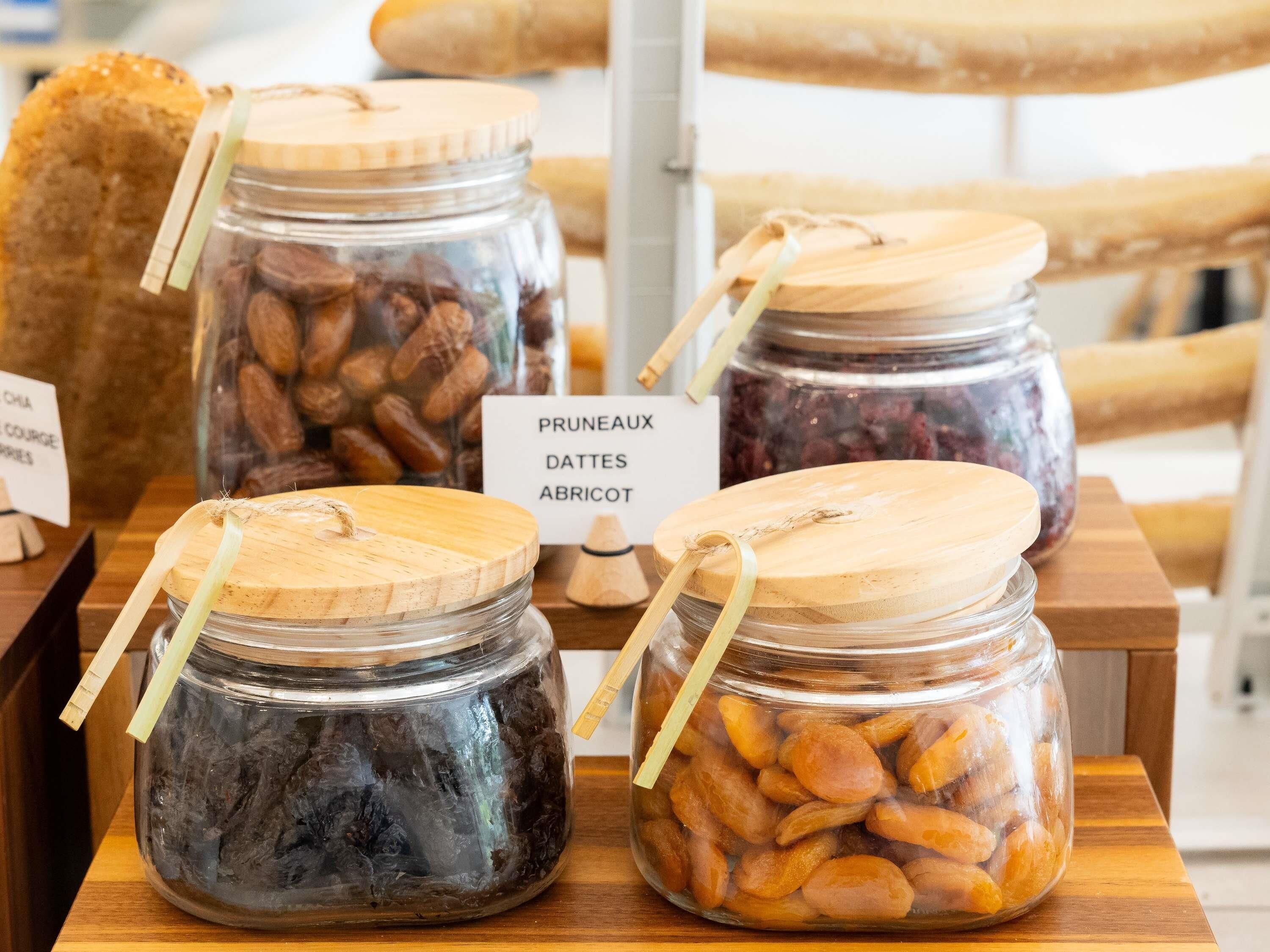 Four glass jars with wooden lids on a wooden table, labeled with different types of dried fruits.