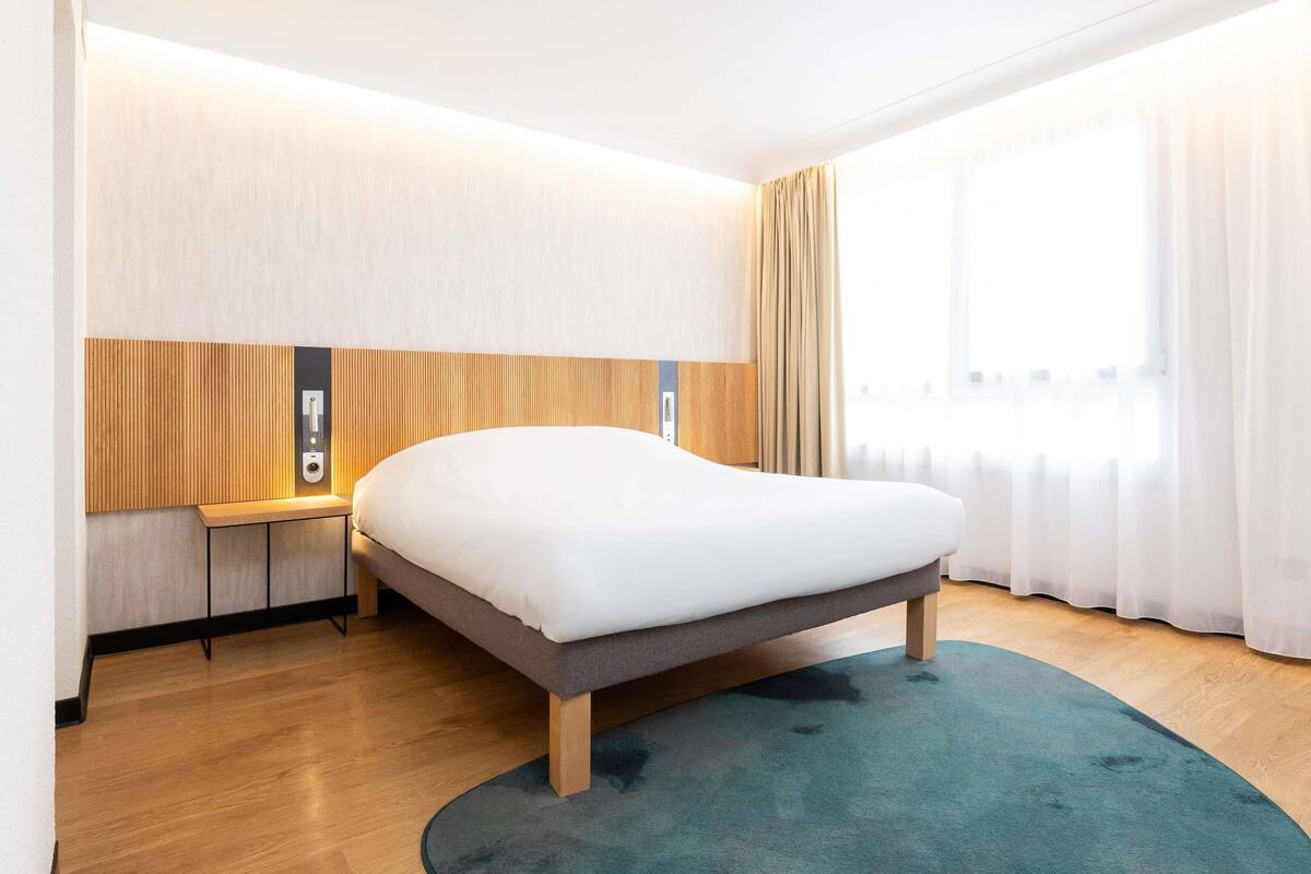 A modern hotel room with a large bed, a bedside table, a built-in TV, and a window with curtains.