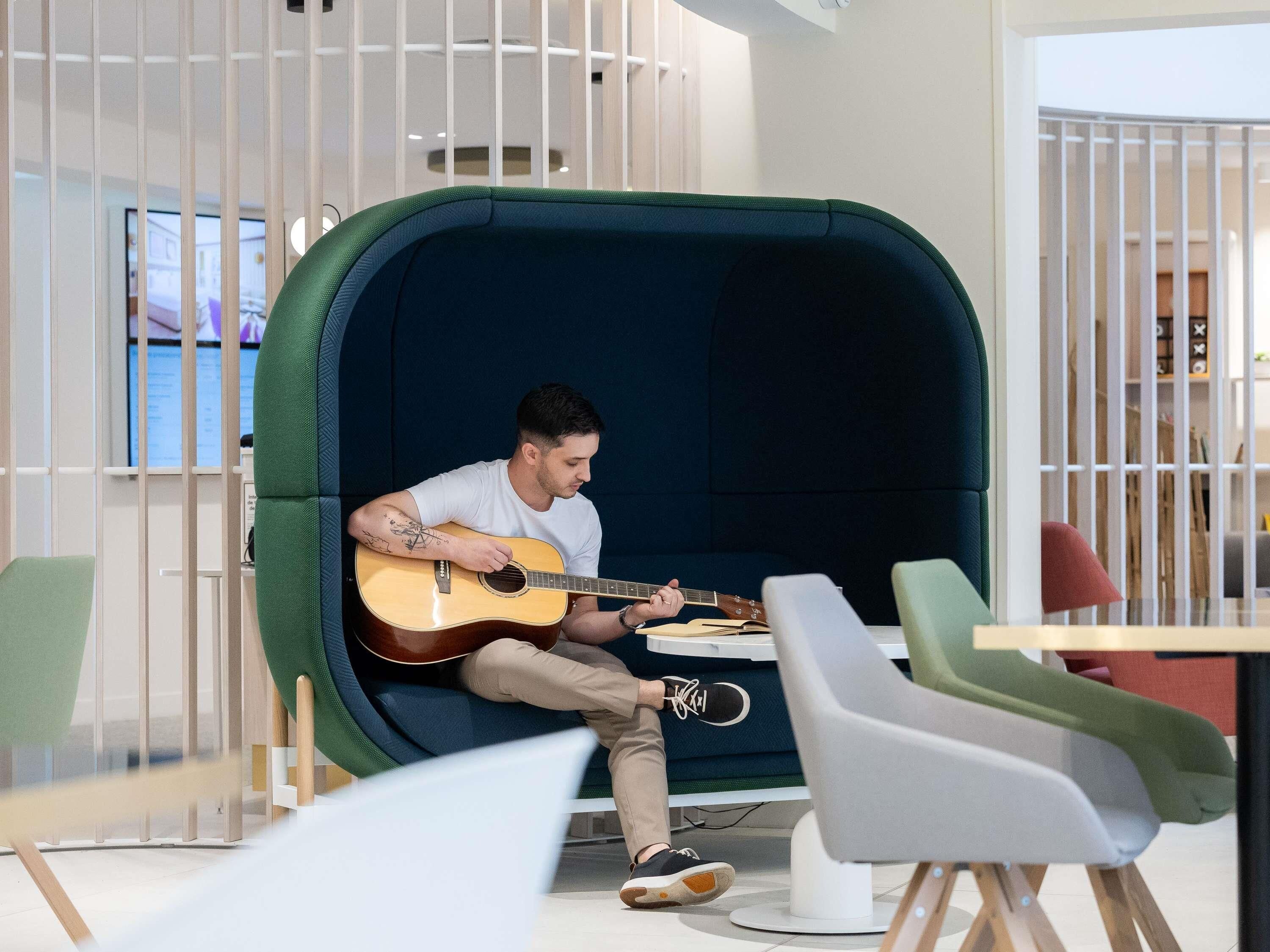 En person spelar akustisk gitarr i en mysig liten vrå med mörkblå inredning, omgiven av moderna möbler och ett vitt räcke.
