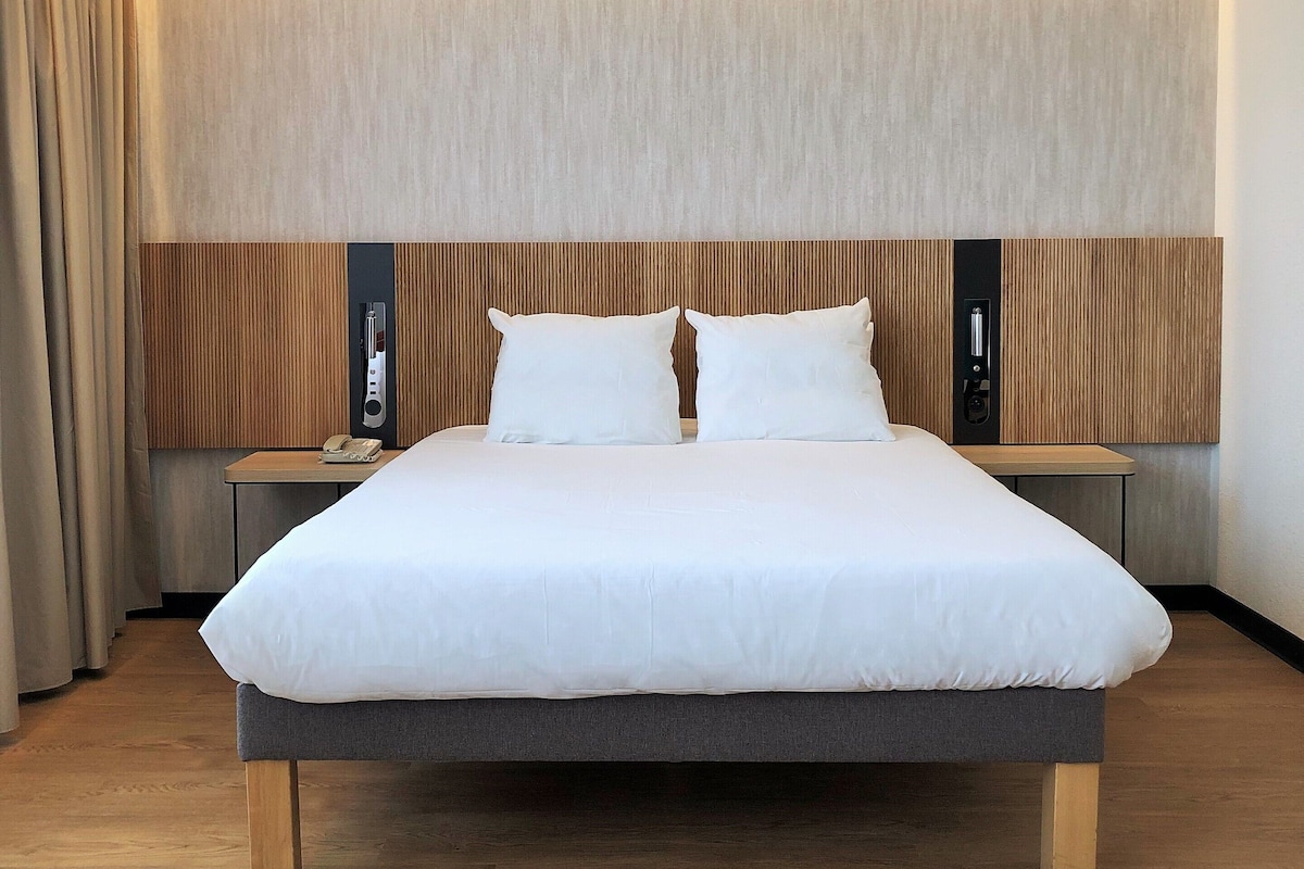 Una habitación de hotel moderna con una cama grande, cabecera de madera, mesitas de noche y un teléfono montado en la pared.