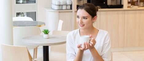 Una persona sentada a una mesa redonda con una taza blanca, un muffin y un vaso de jugo de naranja. Hay un tazón de cereal y una pequeña planta en maceta sobre la mesa.