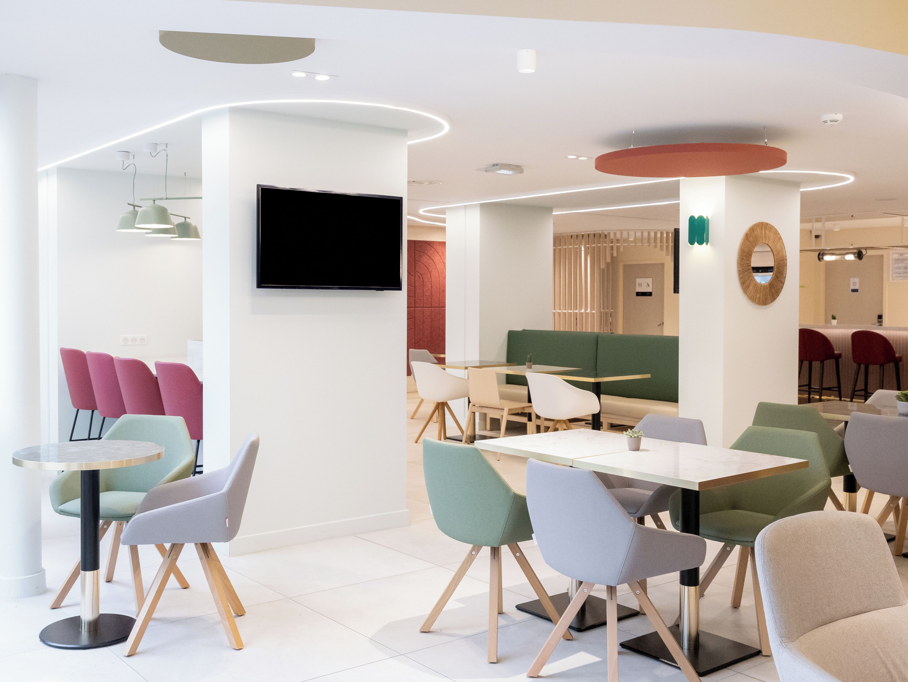 A modern dining area with round tables and chairs, a flat-screen TV mounted on the wall, and a green sofa in the background.
