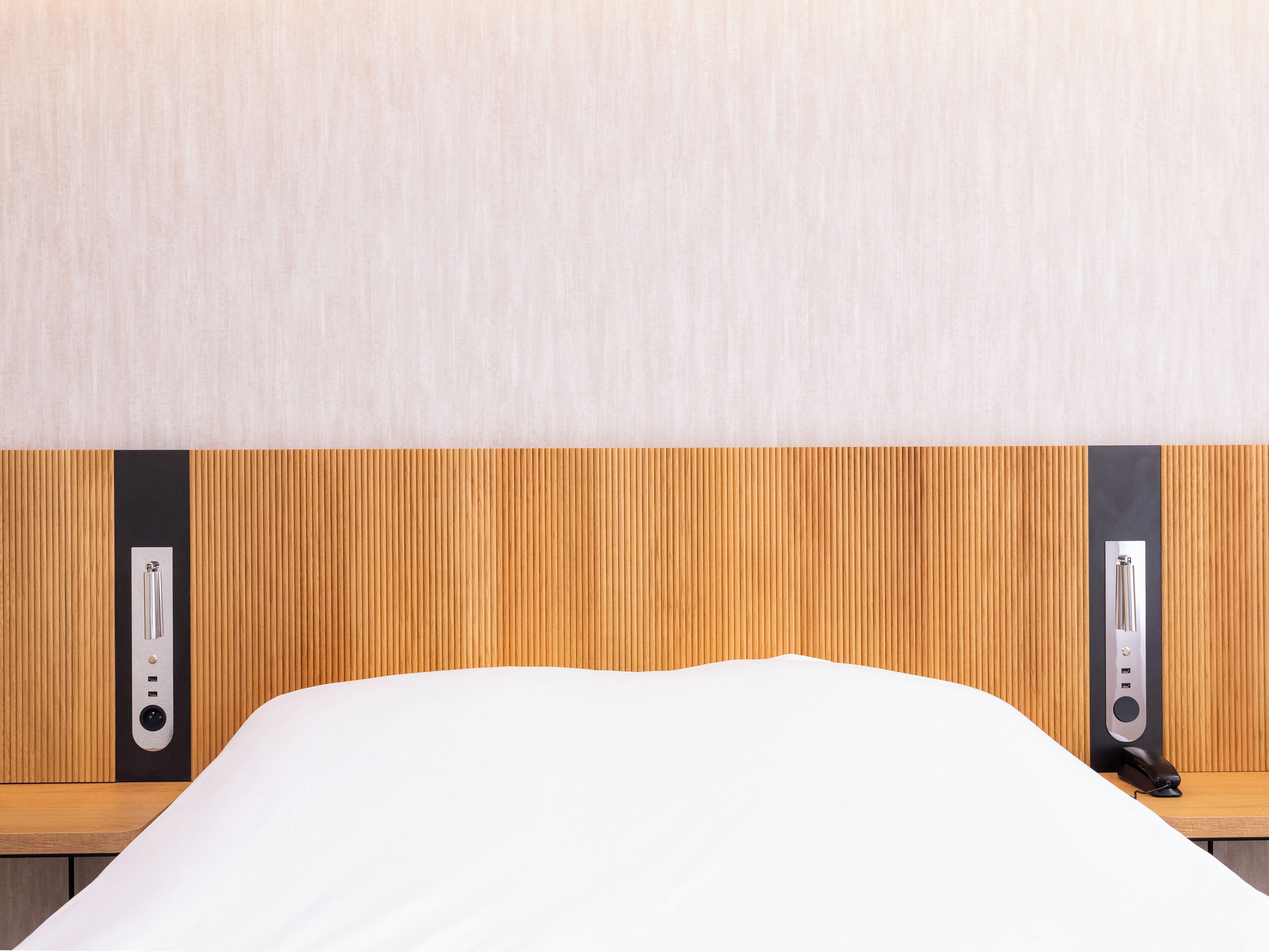Ein Hotelzimmer mit einem hölzernen Kopfteil, einem weißen Bett und einer schwarzen Ladestation mit silbernen Akzenten.