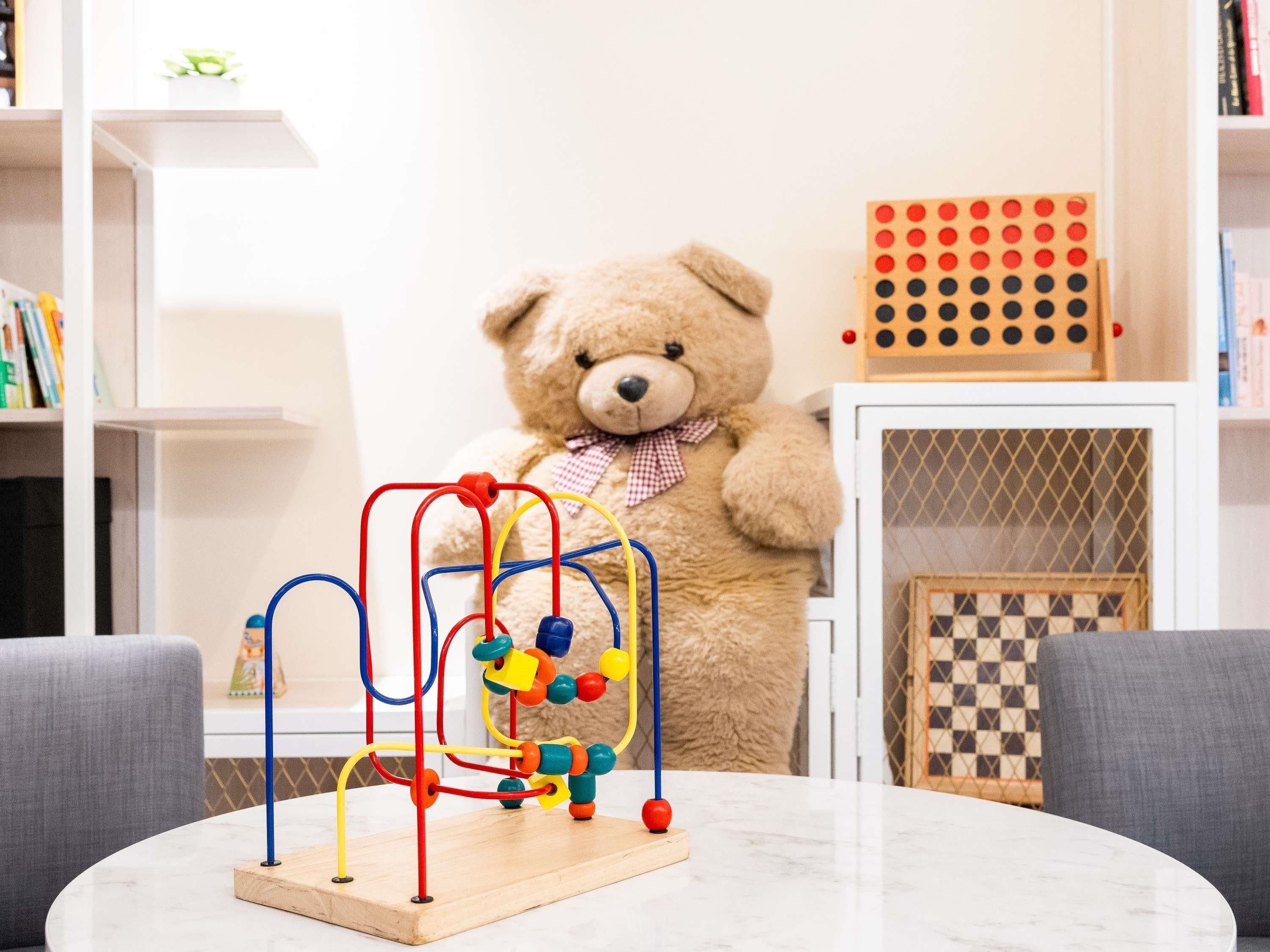 A teddy bear standing behind a wooden play table with a colorful bead maze.