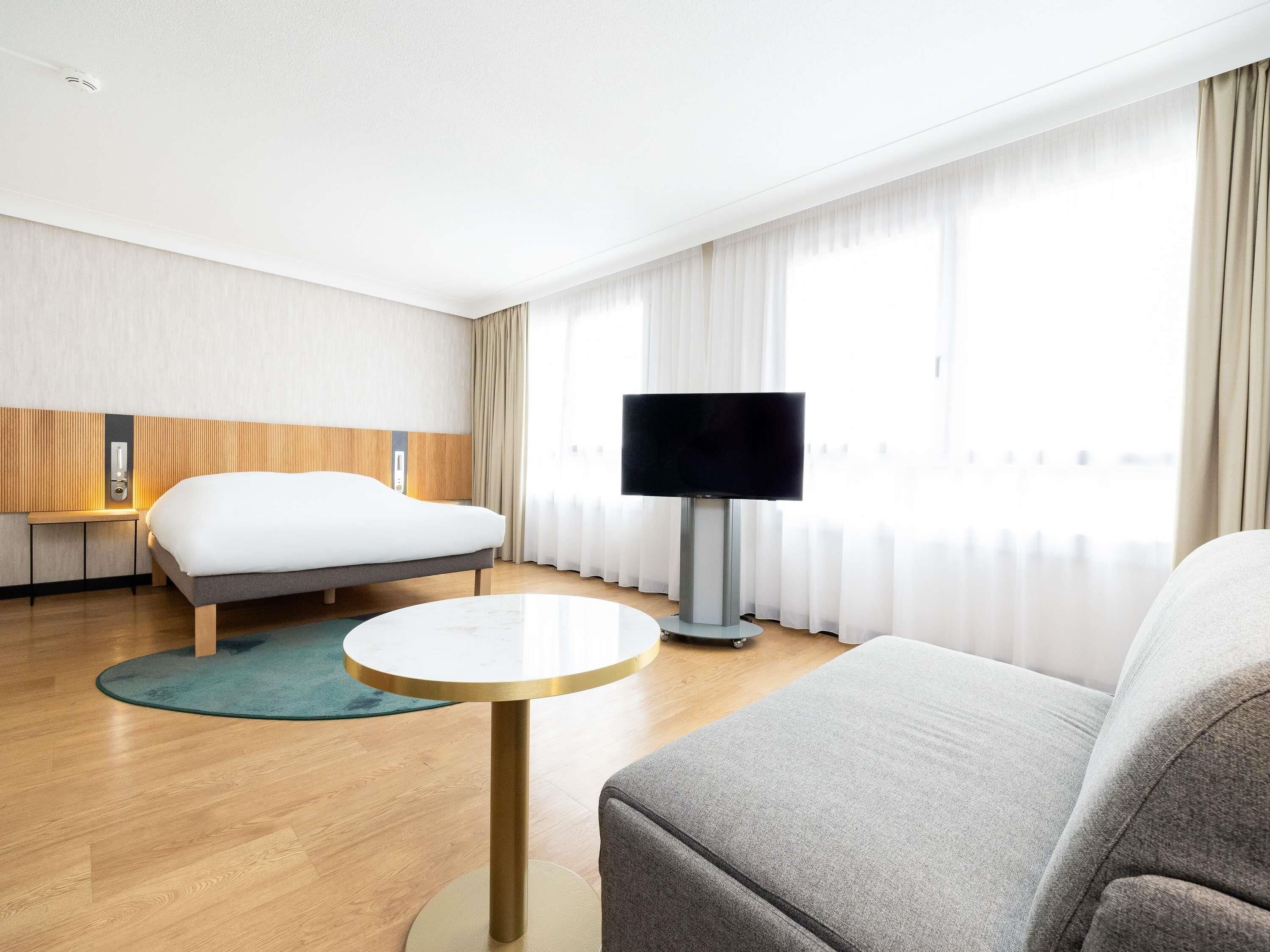 A hotel room with a bed, a TV, a sofa, a small round table, and a window with curtains.