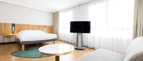 A hotel room with a bed, a TV, a sofa, a small round table, and a window with curtains.