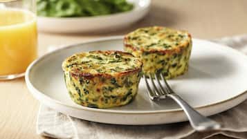 Dos quiches en un plato, un tenedor, un vaso de zumo de naranja y un bol de verduras al fondo.