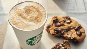A Starbucks cup with a frothy top, next to a plate with a chocolate chip cookie.