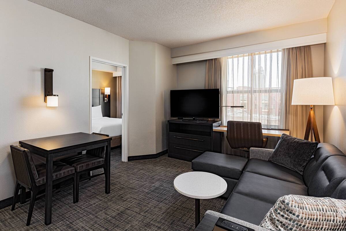 A hotel room with a dark brown sofa, a small round table, a TV, and a desk with a chair.