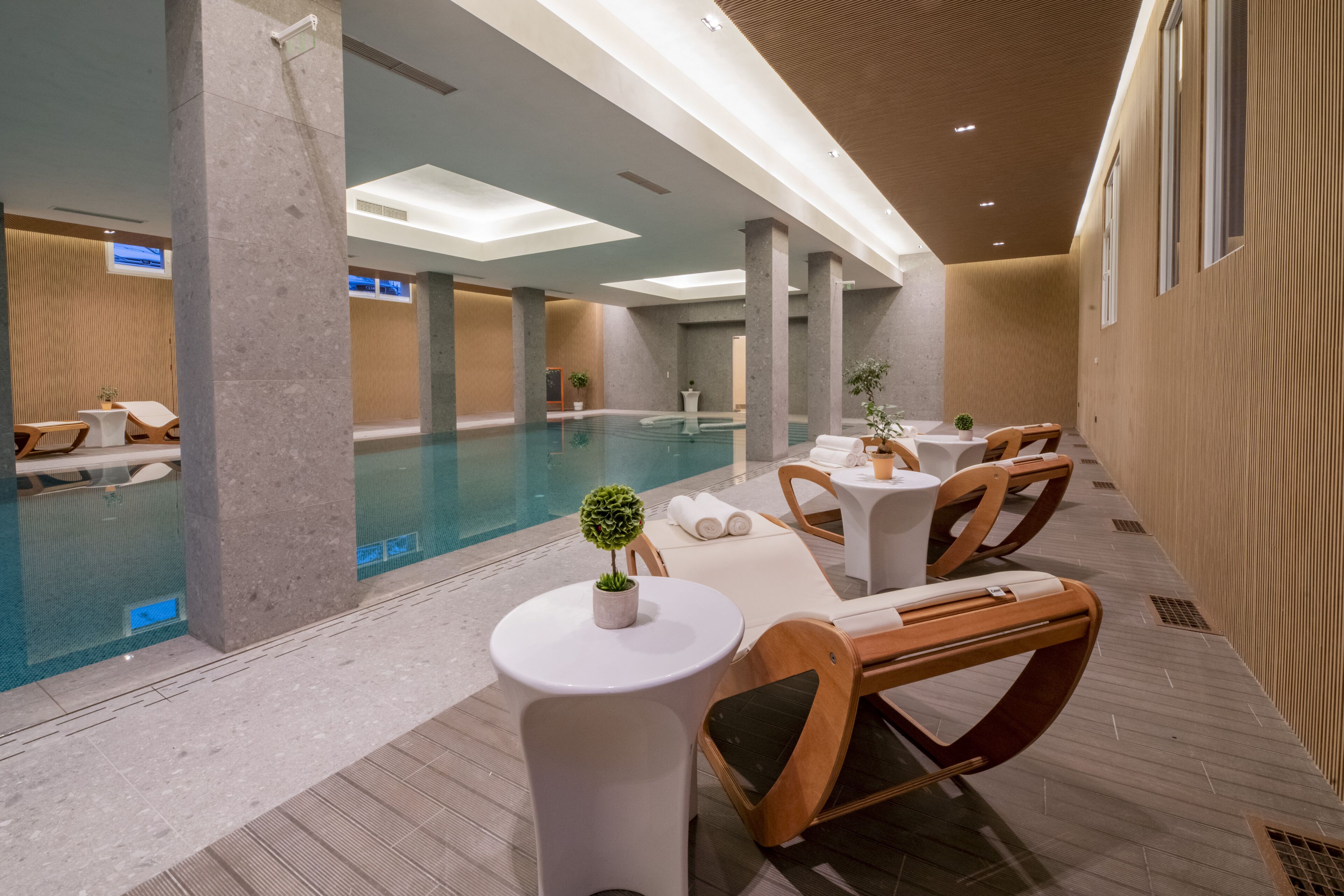 A modern indoor pool area with lounge chairs and tables, a central pool, and a ceiling with recessed lighting.