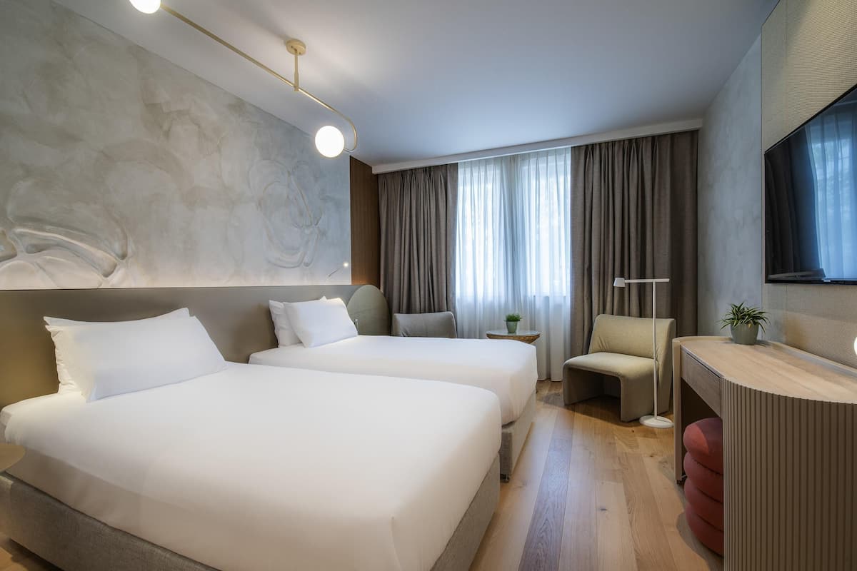 A hotel room with two beds, a desk, a chair, a small table, and a large window with curtains.