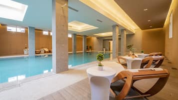 Una piscina interior moderna con sillones y mesas, rodeada de techos altos y luz natural.