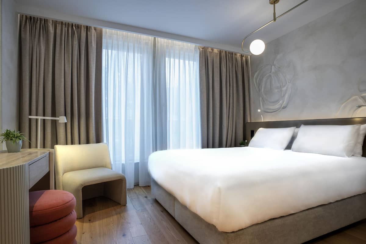 A modern hotel room with a large bed, a desk with a chair, and a small ottoman.