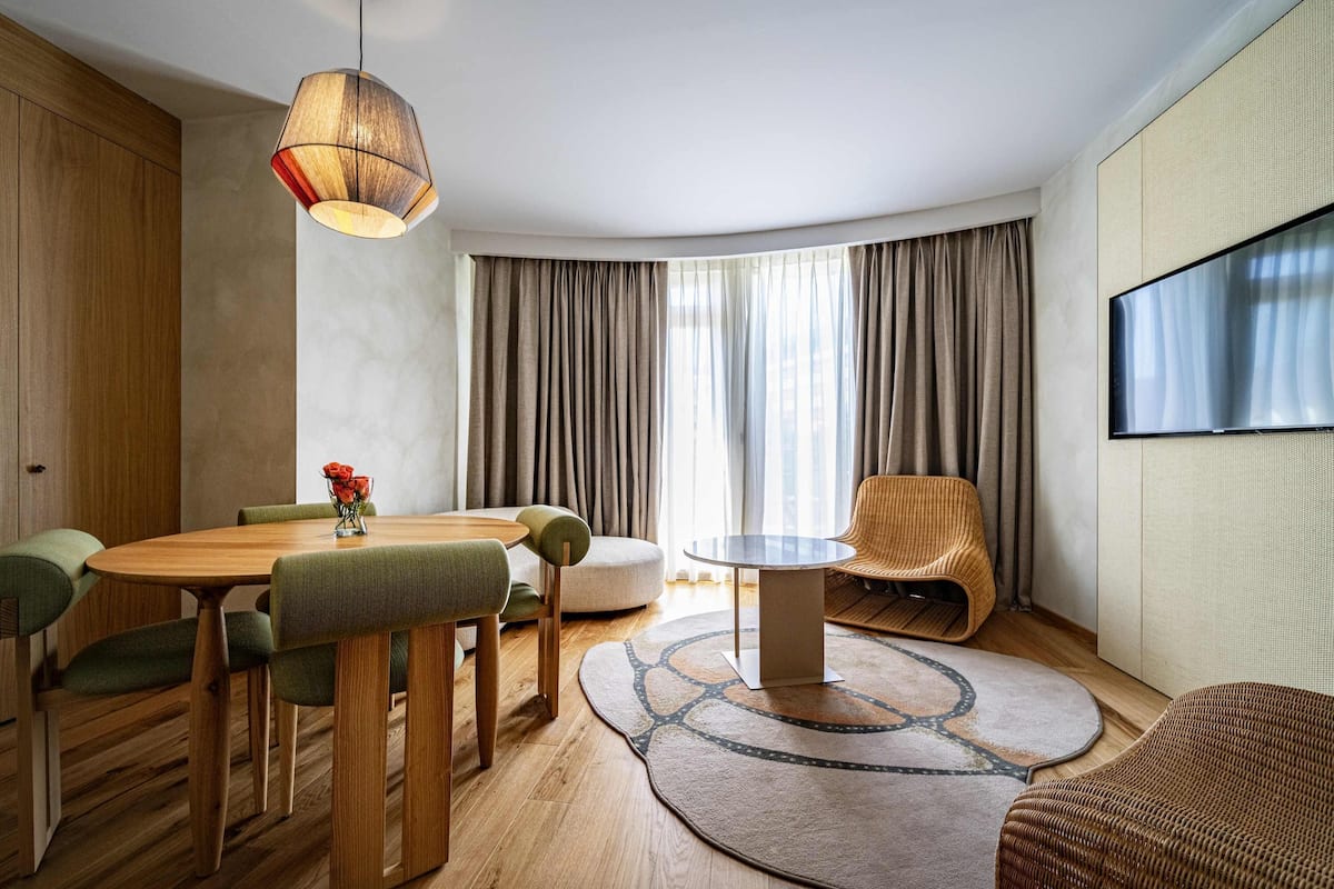 A modern living room with a wooden dining table, green upholstered chairs, a round coffee table, a wicker armchair, and a large TV mounted on the wall.