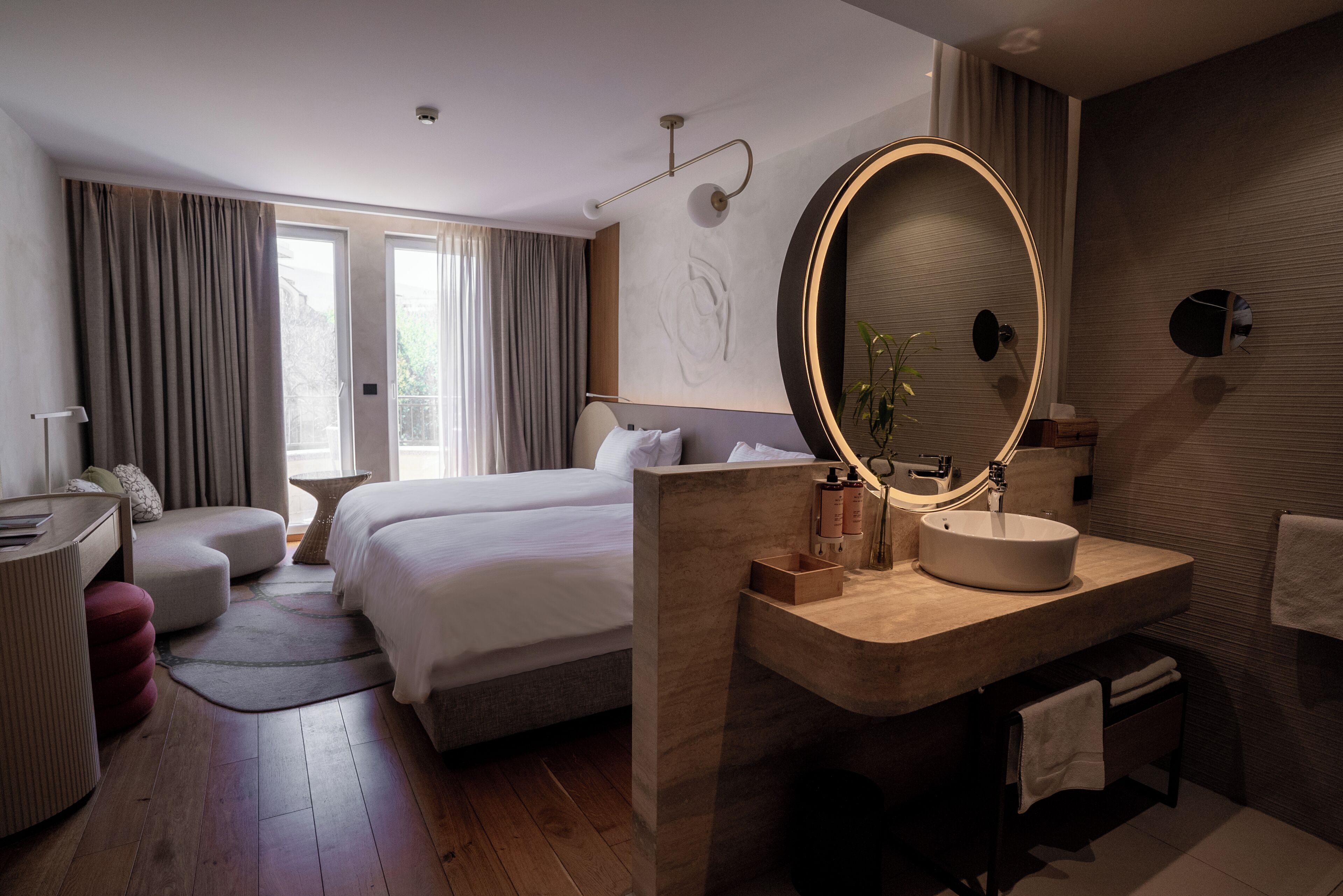 Habitación de hotel con cama, mesita de noche, lámpara y baño con lavabo y espejo.