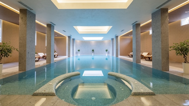 En moderne indendørs swimmingpool med krystalklart vand, omgivet af liggestole og potteplanter.