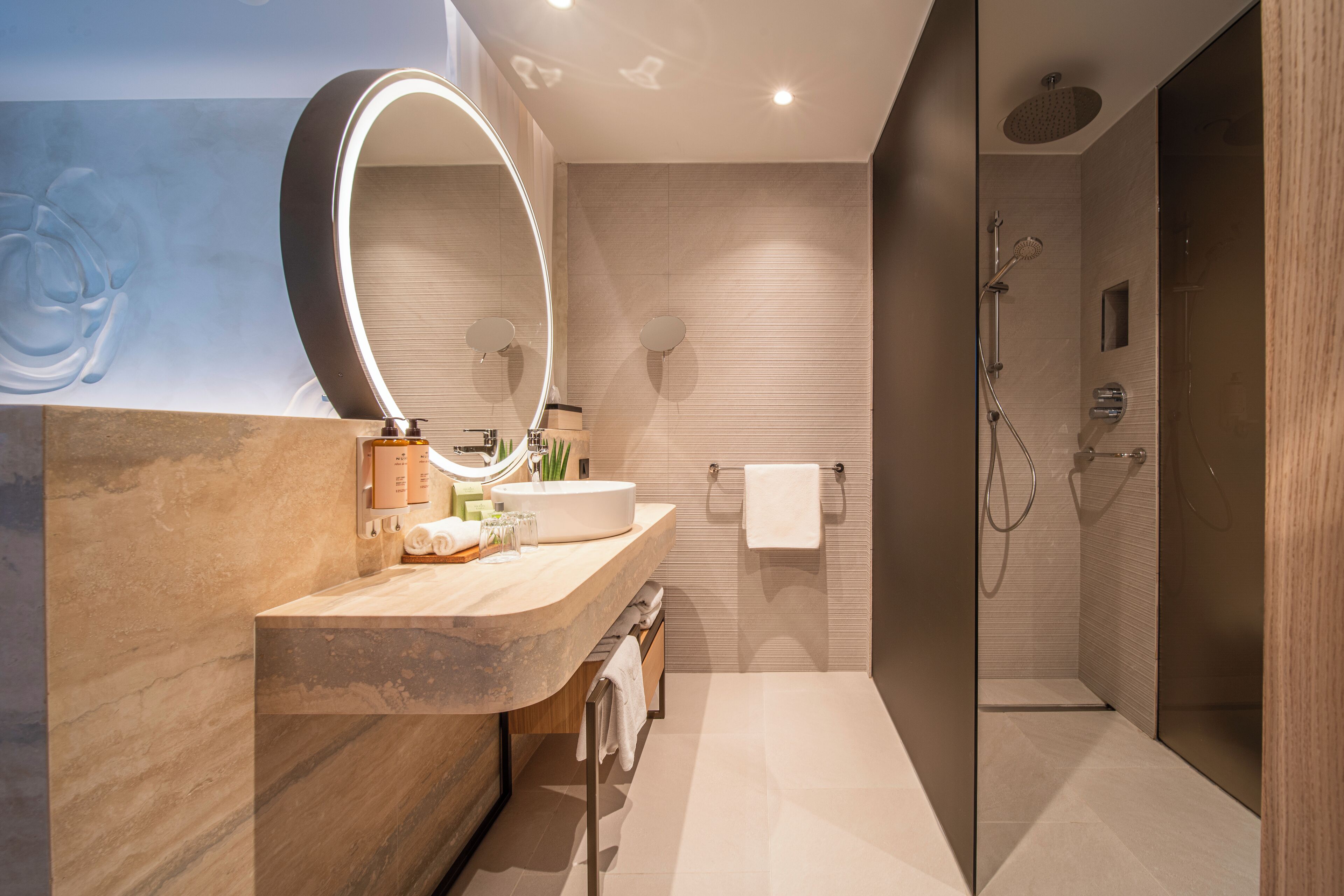 Une salle de bain moderne avec un meuble-lavabo en bois, un grand miroir doté d’un éclairage intégré, une douche avec une porte en verre et un porte-serviettes.