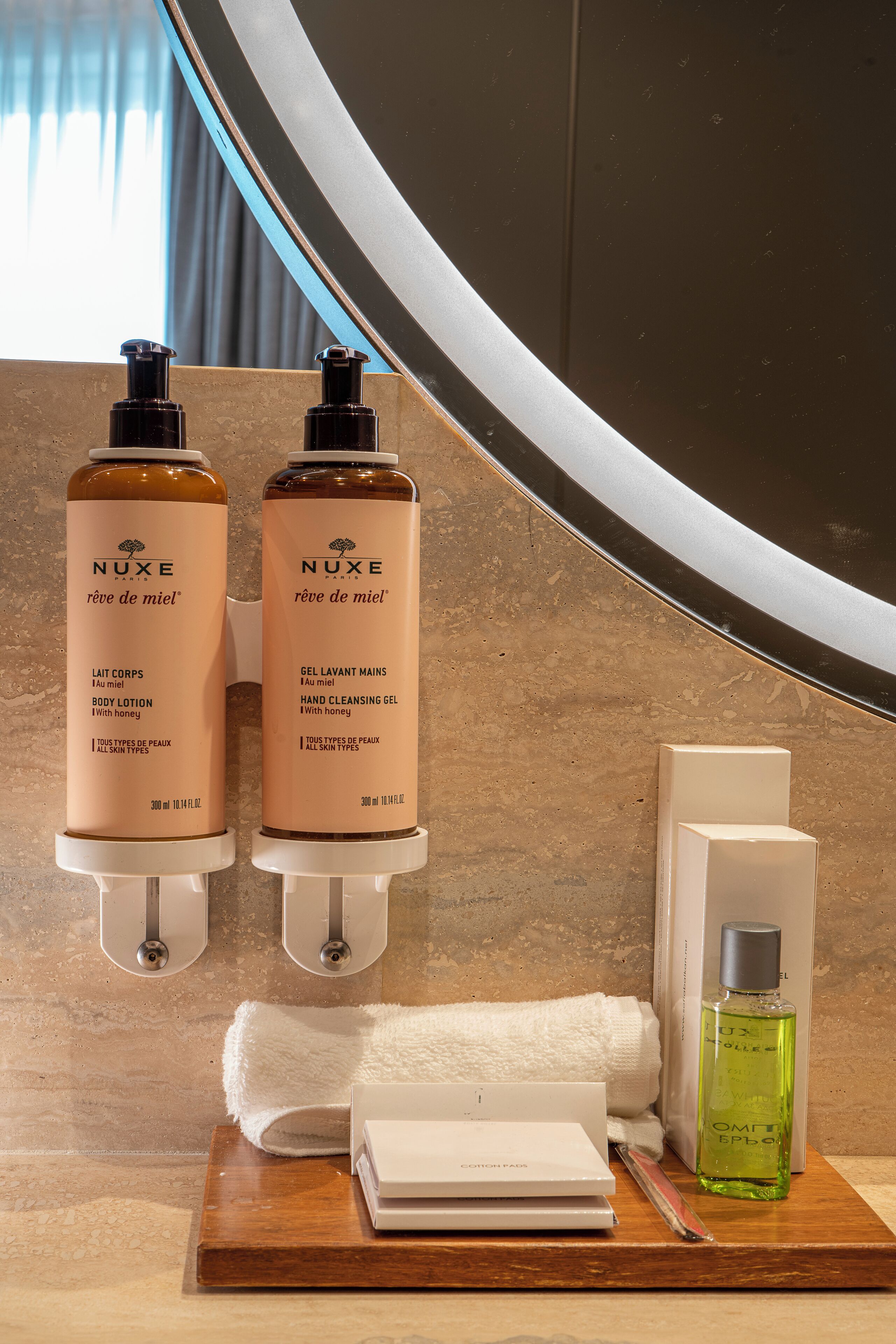 A bathroom with two Nuxe body lotion dispensers, a hand cleansing gel dispenser, a green Nuxe perfume bottle, a white towel, and a toiletry box on a wooden tray.