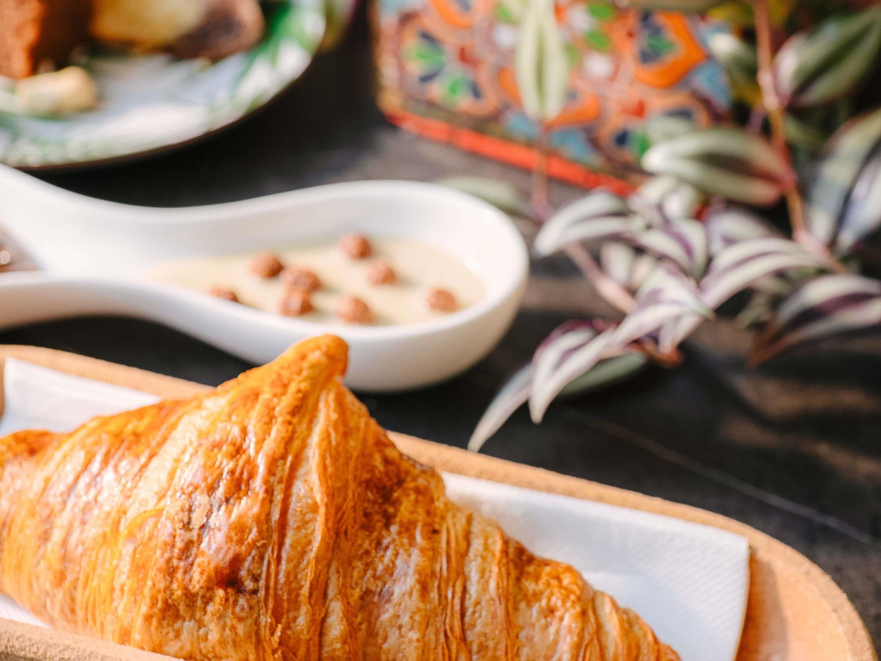Un croissant sobre una tabla de madera con una servilleta blanca, un tazón pequeño y blanco con nueces, y un recipiente colorido con estampado al fondo.