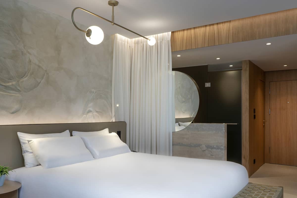 A modern bedroom with a large bed, a bedside table with a plant, a wall-mounted mirror, and a wooden wardrobe.