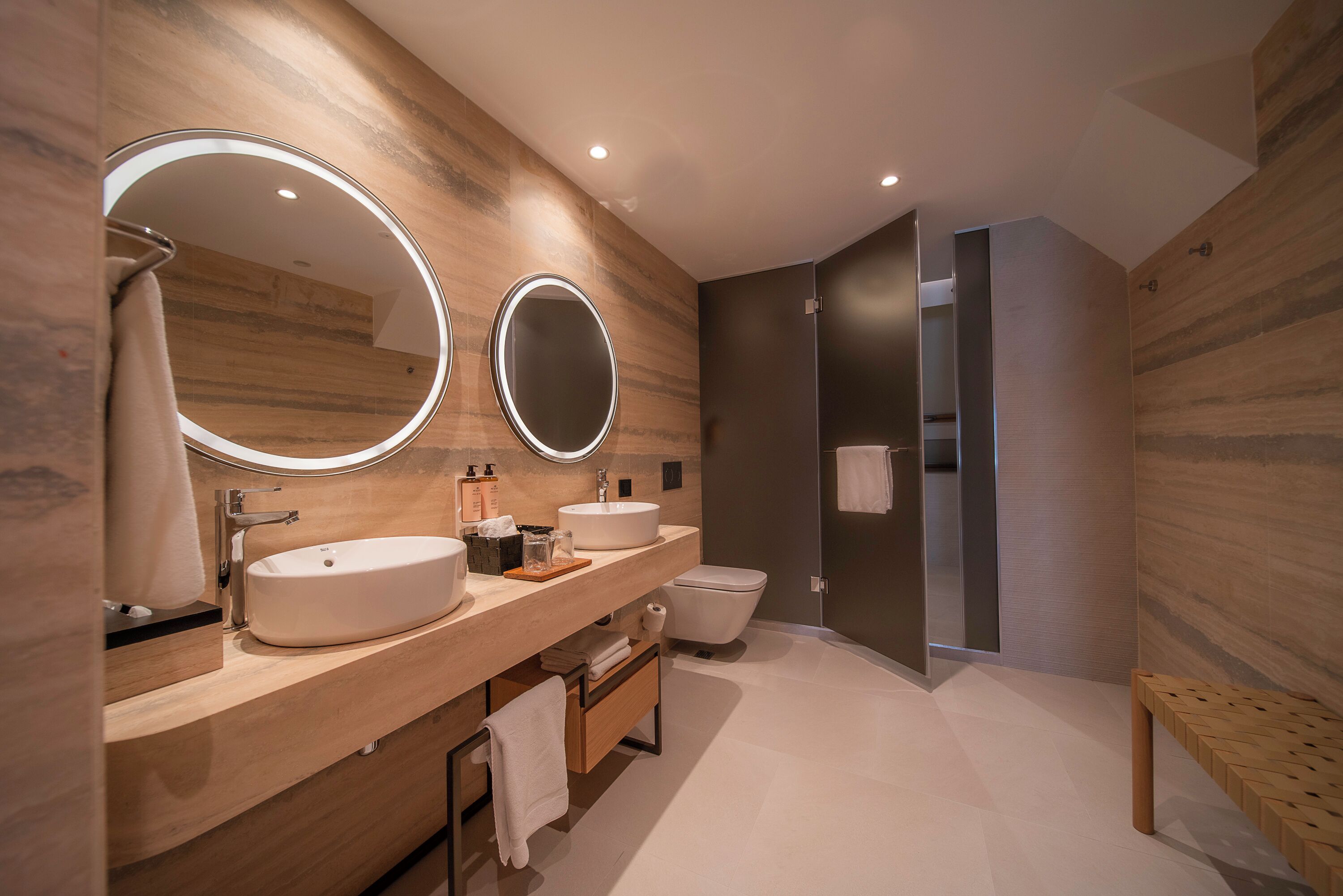 A modern bathroom with a wooden vanity, two round mirrors, a large sink, a toilet, and a shower area.