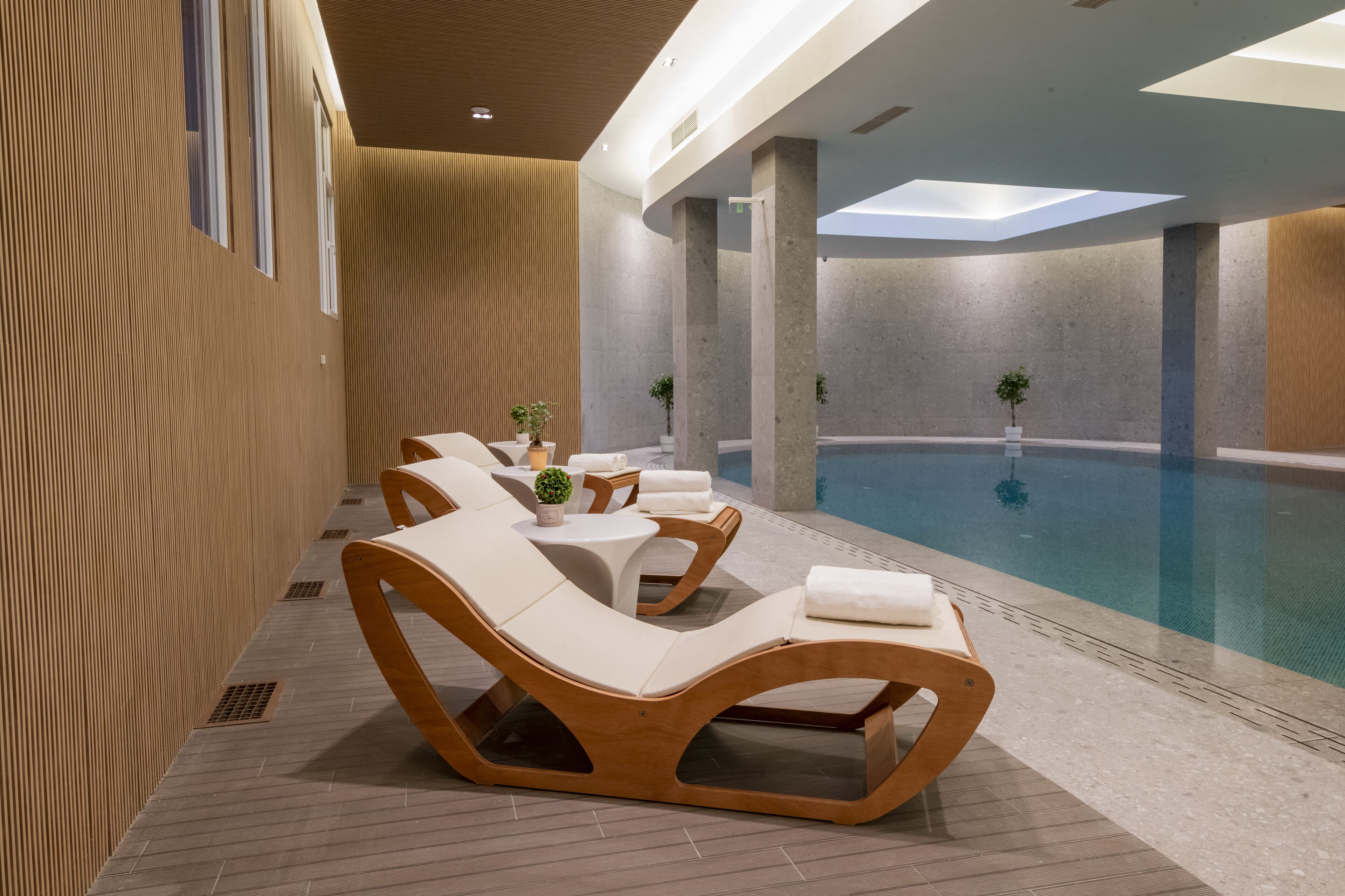 A modern indoor pool area with lounge chairs and small tables, surrounded by wooden paneling and a clear view of the pool.